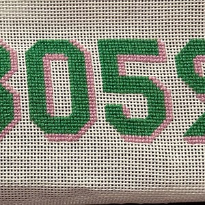 Könnte beinhalten: Eine Stickleinwand mit den Buchstaben BOS in Blockschrift. Die Buchstaben sind mit grünem Garn gestickt und mit einem hellrosa Rand versehen, vor einem weißen Gitterhintergrund. Die Stickerei erzeugt eine texturierte Optik auf dem Stoff.