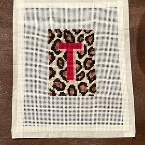 Puede incluir: Lienzo de punto de aguja con un diseño de estampado de leopardo y la letra "T" roja y rosa. El lienzo cuadrado tiene un borde blanco y una cuadrícula de pequeños agujeros para el trabajo de aguja. El diseño está centrado en el lienzo.