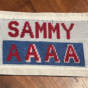 May include: Needlepoint canvas with the name "SAMMY" in red block letters above a blue rectangle with the letters "AAAA" in red. The canvas is white with a grid pattern.