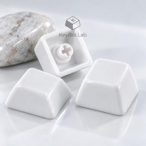 May include: Three glossy white mechanical keyboard keycaps with a smooth, square-shaped surface. One keycap is flipped over to reveal the cross-shaped stem mount on the underside. The text KeyBoi.Lab is printed near the center of the image.
