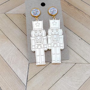 May include: A pair of gold-toned earrings featuring a Christmas theme. Each earring has a small circular top with the text "NUTS ABOUT CHRISTMAS" and a detailed illustration. Below, a white nutcracker soldier design hangs, adding a festive touch.