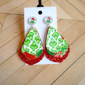 May include: Green and red layered dangle earrings with a Grinch face cameo. The earrings are made of leather and glitter.