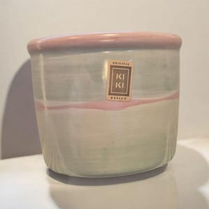 Op de afbeelding: Een lichtgroene en roze keramische plantenpot met een bruin label met de tekst "Original Kiki Design".