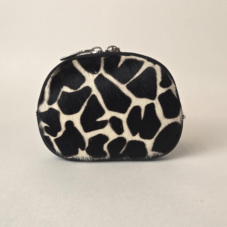 May include: A small, rounded coin purse featuring a textured black and white animal print pattern. The exterior is covered in short, fine hair, and the top edge is secured with a silver-toned metal zipper closure that runs along the curved top.