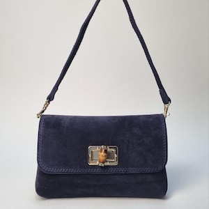 Può includere: Una borsa a mano in camoscio blu navy con forma rettangolare e chiusura a patta. La borsa presenta una chiusura in stile bambù e un cinturino blu navy abbinato. La ferramenta è color oro.