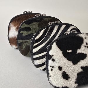 May include: A row of four small, semi-circular zippered pouches made of textured animal-print hair-on hide. The patterns include a black and white cow print, a black and white zebra stripe, a green and black camouflage, and a brown and white hide.