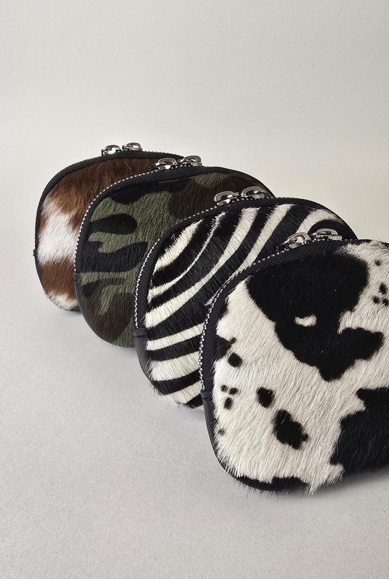 May include: A row of four small, semi-circular zippered pouches made of textured animal-print hair-on hide. The patterns include a black and white cow print, a black and white zebra stripe, a green and black camouflage, and a brown and white hide.
