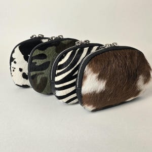 May include: Four small, oval-shaped zippered pouches made of textured hair-on hide. The patterns include a black and white cow print, a green and brown camouflage print, a black and white zebra stripe, and a brown and white cow print.