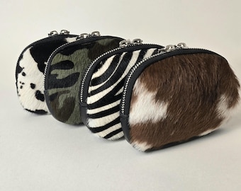 May include: Four small, oval-shaped zippered pouches made of textured hair-on hide. The patterns include a black and white cow print, a green and brown camouflage print, a black and white zebra stripe, and a brown and white cow print.