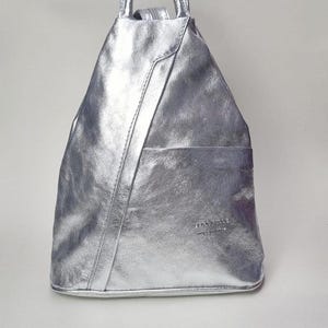 May include: Silver backpack with a top handle and a front pocket. The backpack is made of a shiny, textured material. The bag has a triangular shape and a small handle at the top.