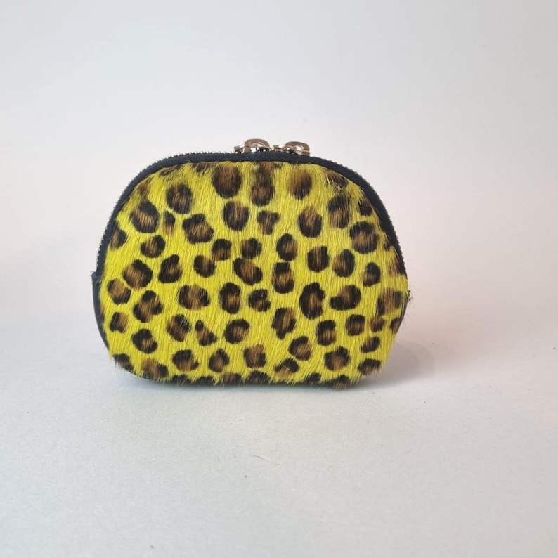 May include: A small, semi-circular coin purse featuring a vibrant yellow and black leopard print pattern on a textured, hair-on-hide material. The pouch is secured with a black zipper closure along the top edge, featuring metallic hardware.