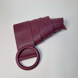 May include: A rolled-up, burgundy leather belt with a matching circular buckle. The belt is made of textured leather and is displayed against a white background. The belt is a fashion accessory.