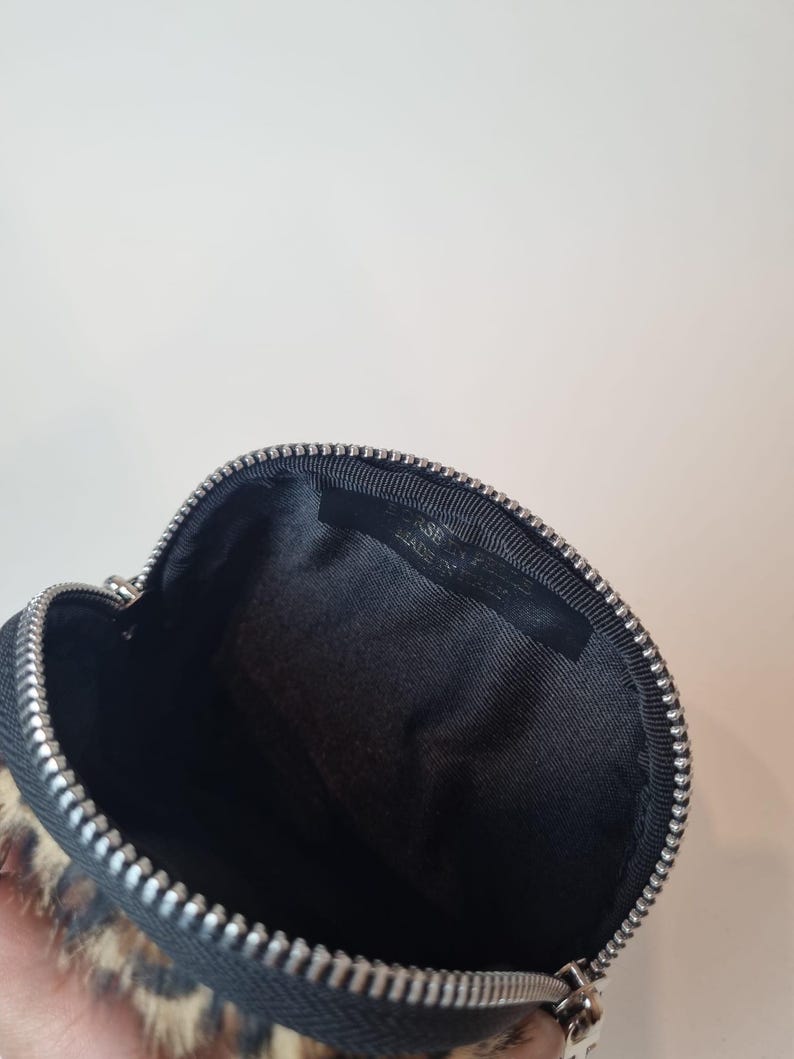 May include: A small, round, zippered pouch featuring a leopard print pattern on the exterior. The interior is lined with black fabric and includes a black tag with the text BORSE IN PELLE MADE IN ITALY stitched inside the top edge.