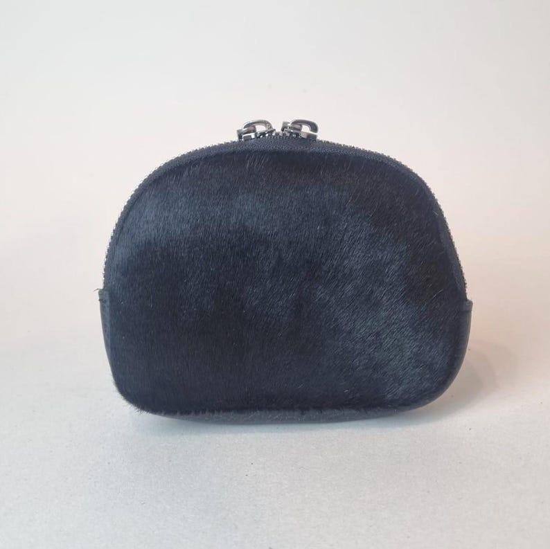 May include: A semi-circular pouch crafted from black hair-on hide leather. The accessory features a smooth, dark texture and is secured at the top with a silver-toned metal zipper closure that runs along the curved edge of the small storage item.