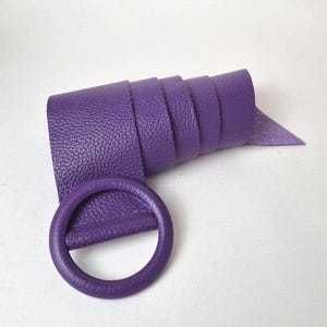 May include: A rolled-up, textured, purple belt with a matching circular buckle. The belt is made of a flexible material and is a solid, vibrant shade of purple. The buckle is a simple, round design.