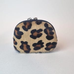 May include: A small, semi-circular coin purse featuring a textured, tan and brown leopard print pattern. The pouch is constructed with a dark trim along the top edge and includes two small, silver-toned metal zipper pulls at the center closure.