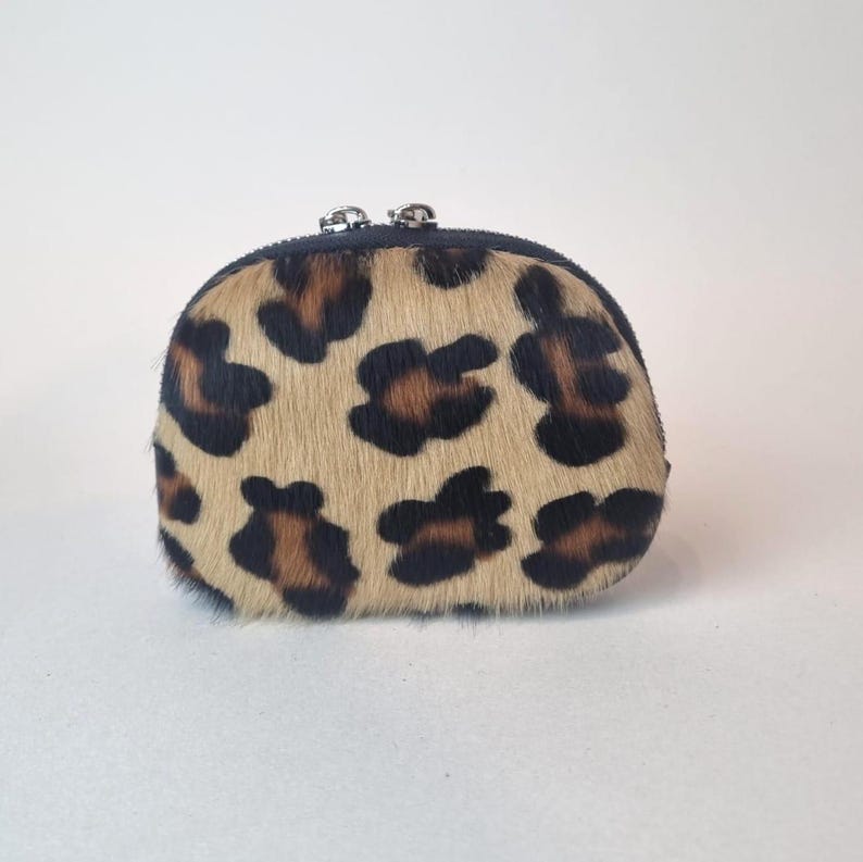 May include: A small, semi-circular coin purse featuring a textured, tan and brown leopard print pattern. The pouch is constructed with a dark trim along the top edge and includes two small, silver-toned metal zipper pulls at the center closure.