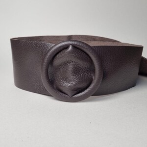 May include: A wide, dark brown leather belt with a circular buckle. The belt is made of textured leather and has a simple, yet elegant design. The buckle is a prominent feature, adding a touch of sophistication to the accessory.