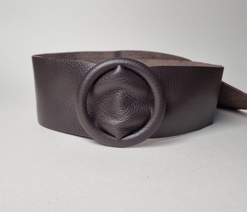 May include: A wide, dark brown leather belt with a circular buckle. The belt is made of textured leather and has a simple, yet elegant design. The buckle is a prominent feature, adding a touch of sophistication to the accessory.