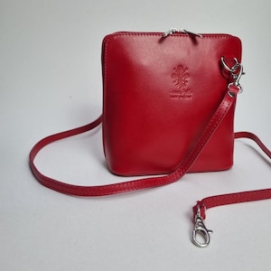 May include: A bright red leather crossbody bag with a silver zip and strap fastenings. The bag has a small embossed emblem and the words "made in Italy". The strap is detachable, and the bag is rectangular.