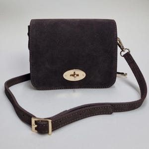 Puede incluir: Bolso bandolera de ante marrón oscuro con cierre y herrajes dorados. El bolso tiene forma rectangular con solapa y una correa larga y ajustable. El bolso está hecho de un material suave y texturizado.