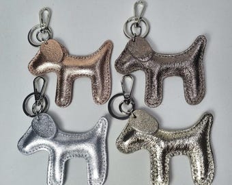 May include: Four metallic dog-shaped keychains in shades of rose gold, bronze, silver, and gold. Each keychain has a silver clasp and ring. The keychains are arranged on a white surface.