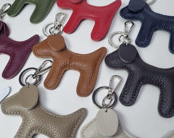 May include: A collection of leather dog-shaped keychains in various colors, including green, red, brown, and gray. Each keychain features a silver-colored clasp and ring for attaching keys. The keychains are arranged on a white surface.