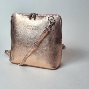 May include: Rose gold crossbody bag with a metallic finish. The bag has a rectangular shape with a zip closure and a detachable strap. The text "VERA PELLE MADE IN ITALY" is visible on the front.