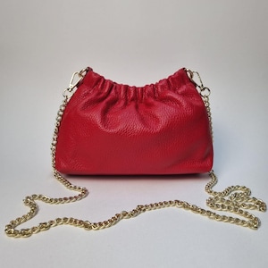 May include: A bright red leather crossbody bag with a gold chain strap. The bag features a gathered top and a textured finish. The gold chain is attached with gold-coloured clasps.
