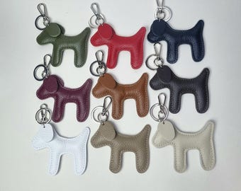 May include: A collection of nine leather dog-shaped keychains in various colors, including green, red, navy, burgundy, brown, black, white, tan, and cream. Each keychain has a silver-colored clasp.