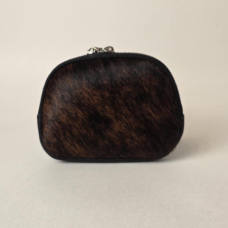May include: A small, semi-circular coin purse featuring a dark brown and black brindle patterned hair-on-hide texture. The pouch is finished with a black leather trim along the edges and a silver-toned metal zipper closure at the top center.
