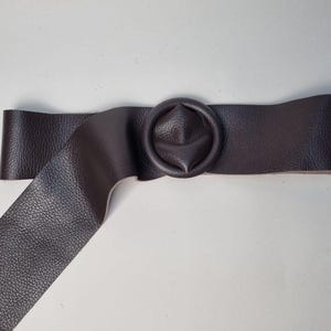 May include: A dark brown leather belt with a circular buckle. The belt has a textured surface and a wide strap. The buckle is a matching dark brown color and has a unique, rounded design. The belt is approximately 2 inches wide.