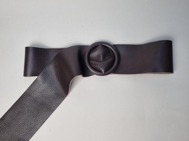 May include: A dark brown leather belt with a circular buckle. The belt has a textured surface and a wide strap. The buckle is a matching dark brown color and has a unique, rounded design. The belt is approximately 2 inches wide.