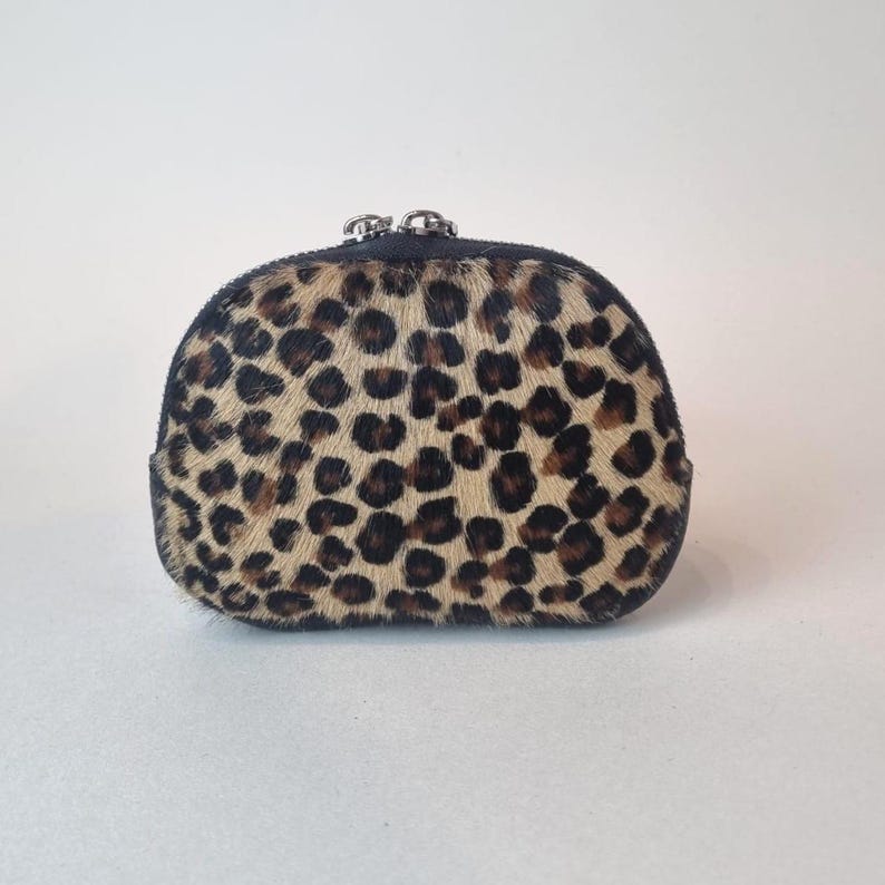 May include: A small, semi-circular coin purse featuring a textured leopard print pattern in shades of tan, brown, and black. The pouch is constructed with a black leather trim and includes a silver-toned metal zipper closure at the top.