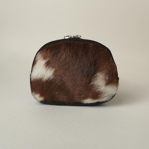 May include: A small, semi-circular coin purse featuring a textured cowhide exterior with a pattern of brown and white patches. The pouch is finished with a black fabric trim along the edges and a silver-toned metal zipper closure at the top.