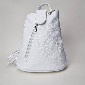 Puede incluir: Una mochila de cuero blanco con asa superior y cremallera diagonal. La bolsa tiene un cierre de solapa con un cierre de metal plateado. Las correas de la mochila son visibles en el lado izquierdo.