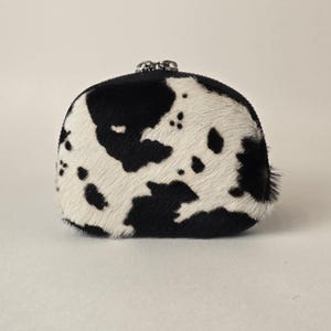 May include: A small, rounded coin purse featuring a textured cowhide pattern with irregular black and white patches. The accessory has a silver-toned metal kiss-lock closure at the top, providing a secure opening for the main compartment.
