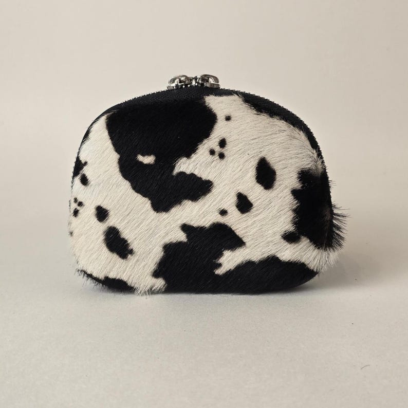 May include: A small, rounded coin purse featuring a textured cowhide pattern with irregular black and white patches. The accessory has a silver-toned metal kiss-lock closure at the top, providing a secure opening for the main compartment.