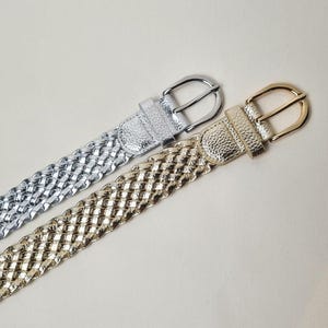 May include: Two braided belts with silver and gold buckles. The belts are made of a woven material and have a metallic finish.