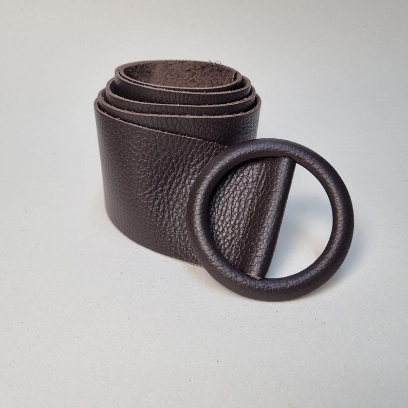 May include: A rolled-up dark brown leather belt with a matching round buckle. The belt has a textured surface and is displayed on a light background. The buckle is a simple, circular design.
