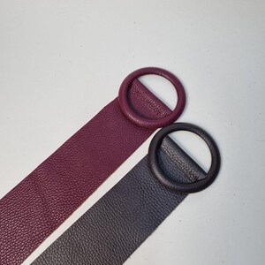 May include: Two leather belts with circular buckles. One belt is a deep burgundy color, and the other is a dark charcoal gray. Both belts have a textured surface and are set against a white background. The buckles are the same color as the belts.
