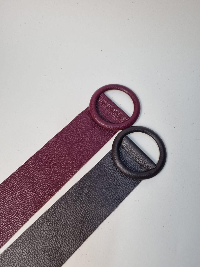 May include: Two leather belts with circular buckles. One belt is a deep burgundy color, and the other is a dark charcoal gray. Both belts have a textured surface and are set against a white background. The buckles are the same color as the belts.