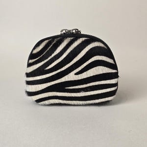 May include: A small, semi-circular coin purse featuring a black and white zebra stripe pattern. The exterior is covered in a textured, hair-on hide material. A silver-toned metal zipper closure is located along the top edge of the pouch.