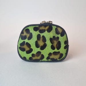 May include: A small, rounded coin purse featuring a vibrant lime green and black leopard print pattern on a textured, hair-on-hide material. The pouch is finished with a dark trim along the edges and a metallic zipper closure at the top.