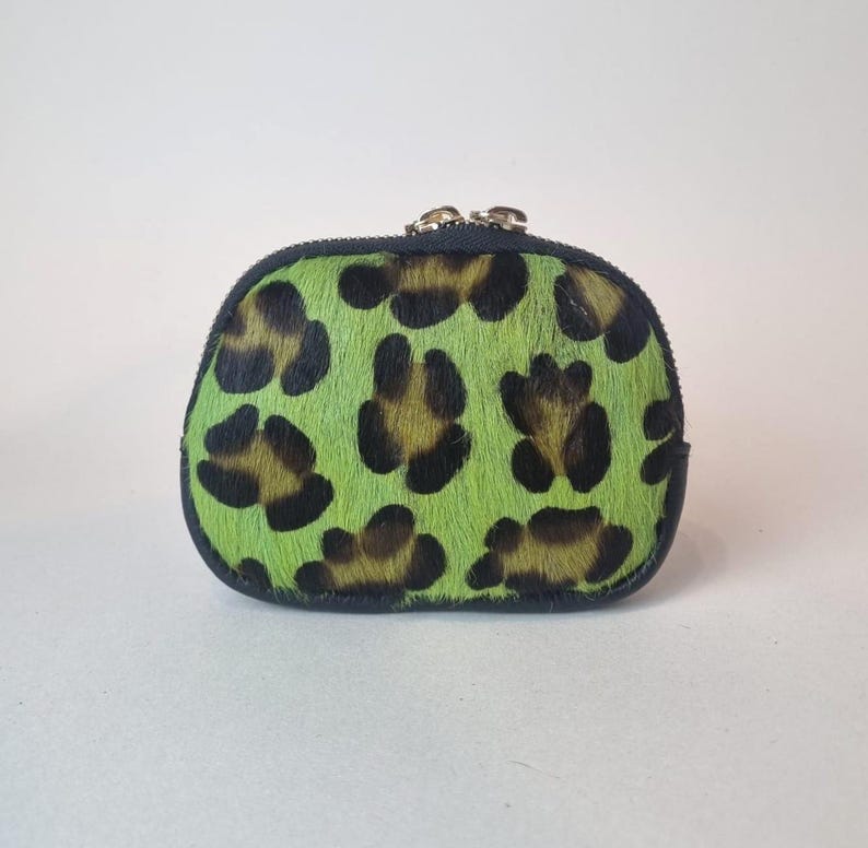 May include: A small, rounded coin purse featuring a vibrant lime green and black leopard print pattern on a textured, hair-on-hide material. The pouch is finished with a dark trim along the edges and a metallic zipper closure at the top.