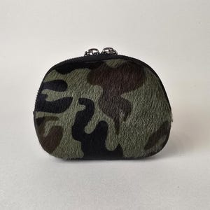 May include: A small, rounded coin purse featuring a textured camouflage pattern in shades of olive green, dark brown, and black. The pouch is constructed with a fuzzy, hair-on hide material and is secured at the top with a black zipper closure.