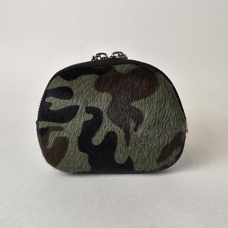May include: A small, rounded coin purse featuring a textured camouflage pattern in shades of olive green, dark brown, and black. The pouch is constructed with a fuzzy, hair-on hide material and is secured at the top with a black zipper closure.