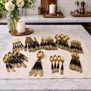 May include: A collection of gold-toned flatware with black handles and ornate detailing. The set includes forks, knives, spoons, and serving spoons, arranged on a neutral-colored linen surface. The flatware is displayed on a marble countertop.