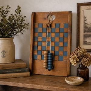May include: A wooden wall-mounted game board featuring a grid of alternating natural wood and blue square tiles. A heart-shaped cutout is at the top, and two vertical strings hang from a central button, each holding a stack of wooden counting beads.