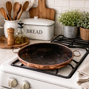 May include: A copper-colored pan with two handles sits on a white stove. The pan has a dark interior and a shiny copper rim. Other kitchen items include wooden spoons, a bread box, and a small potted plant.
